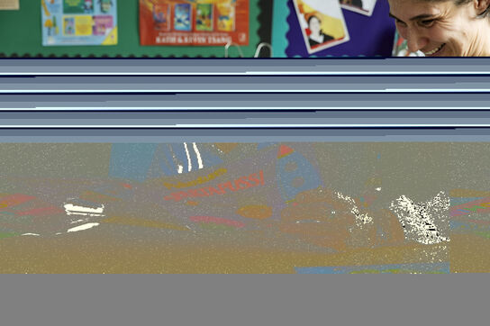 A primary school aged boy reading a storybook in a classroom with his teacher, they are both smiling.