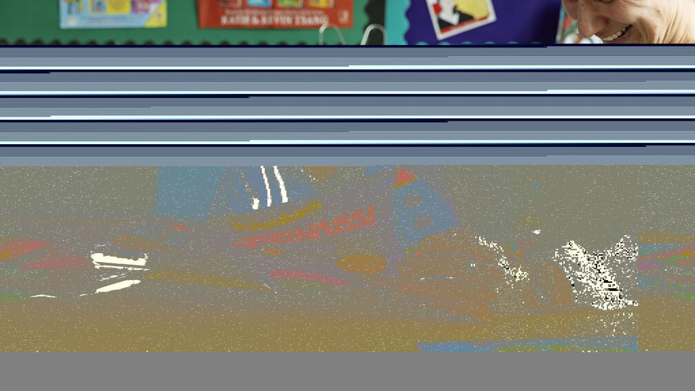 A primary school aged boy reading a storybook in a classroom with his teacher, they are both smiling.