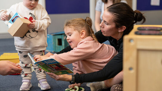 A child and a woman sharing a book - the child is smiling and leaning forward to touch the book. A toddler is walking nearby.
