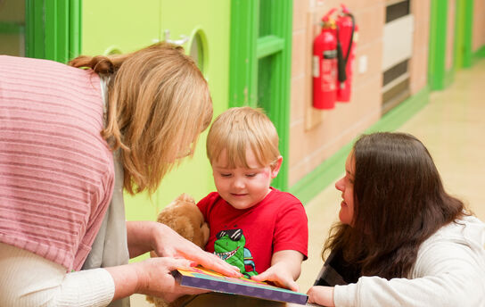 Two early-years practitioners are showing a young boy a book in a nursery setting