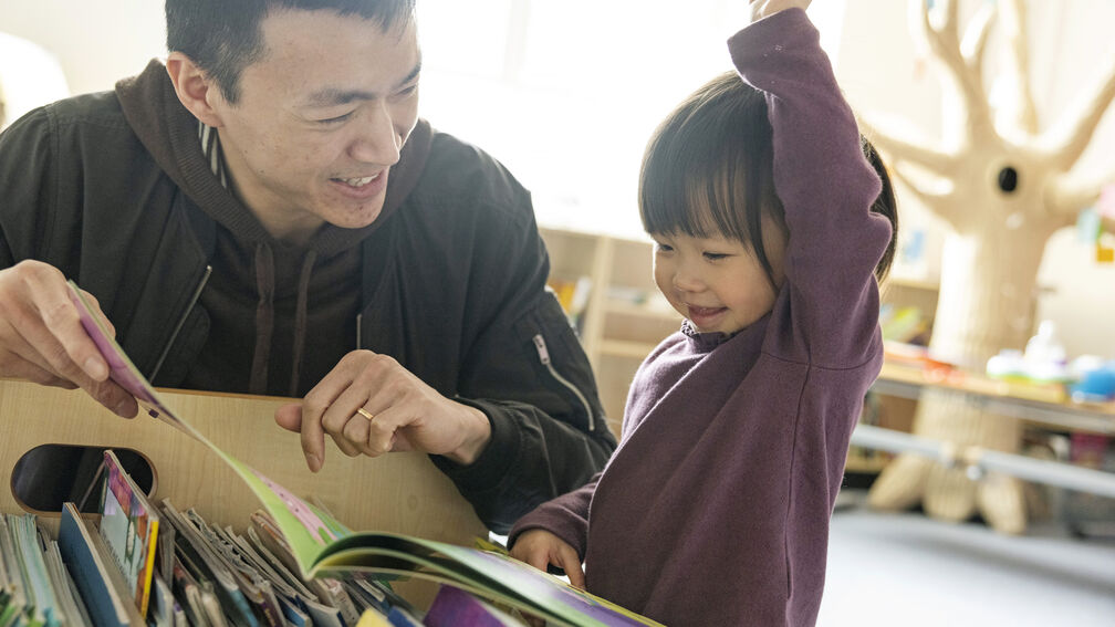 A toddler is choosing books in the library with her dad, she's smiling and waving her hand in the air while she looks at a book and he is smiling at her.