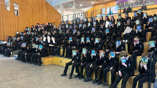 A large group of children in a hall at Oldham Academy holding up their Bookbuzz books in front of their faces
