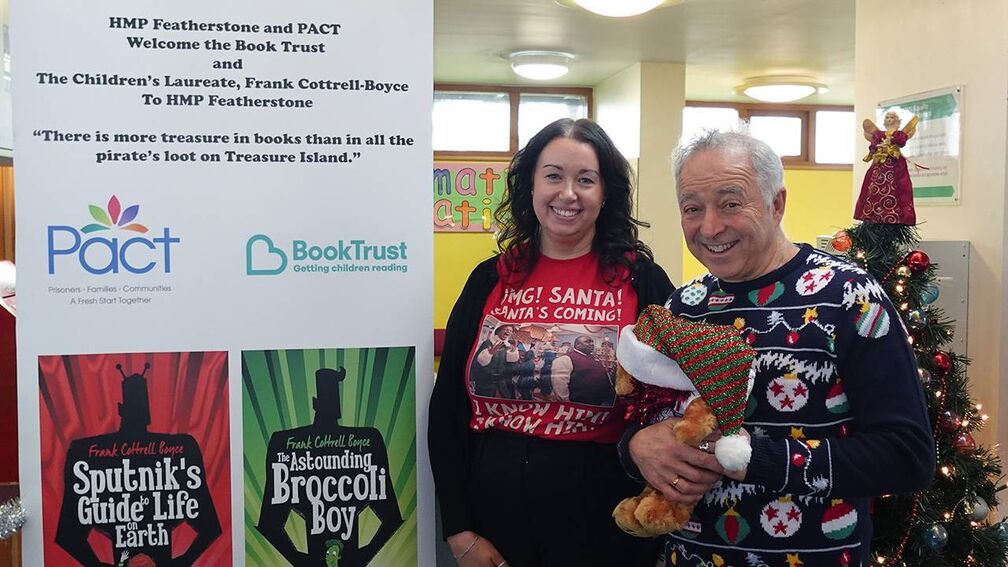 Frank Cottrell-Boyce wearing a Christmas jumper and holding a teddy, arriving at Featherstone prison, next to a banner with the Pact and BookTrust logos on it.