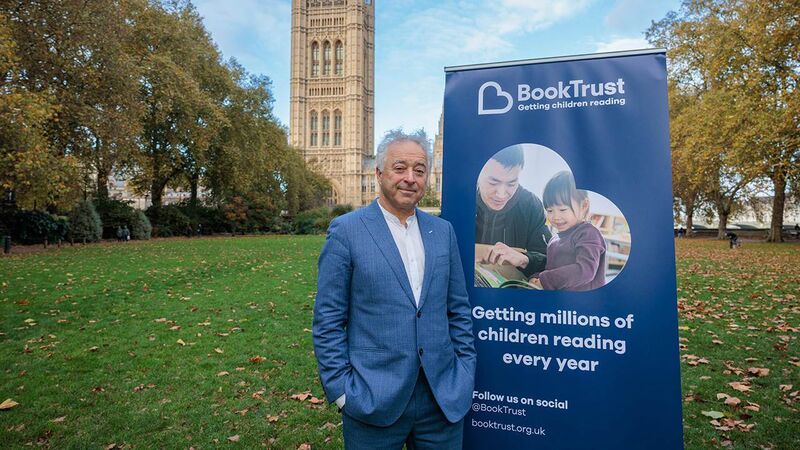 Frank Cottrell-Boyce is standing outside the Houses of Parliament standing in front of a BookTrust banner which says 'Getting millions of children reading every year'