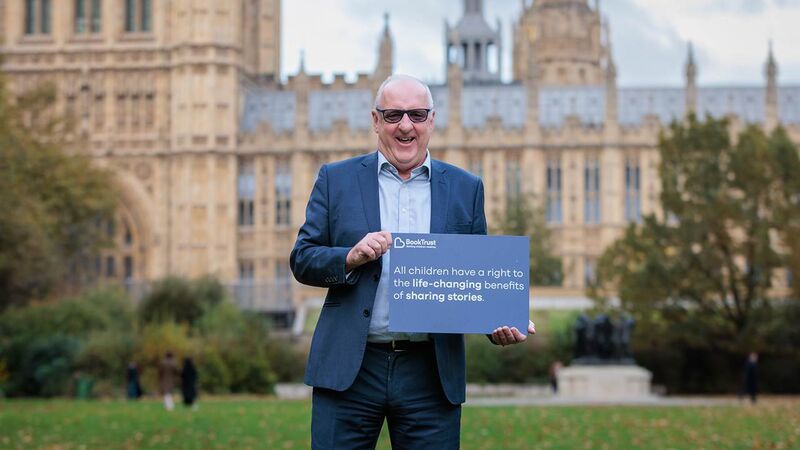 BookTrust Chair of Trustees John Coughlan standing outside the Houses of Parliament, holding a sign that says 'All children have a right to the life-changing benefits of sharing stories.'