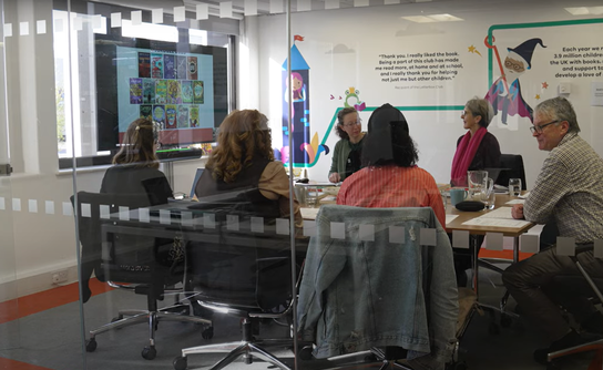 Six adults are sitting around a conference room table, having a group discussion