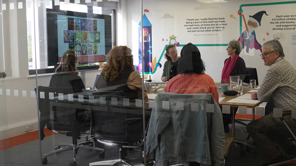 Six adults are sitting around a conference room table, having a group discussion