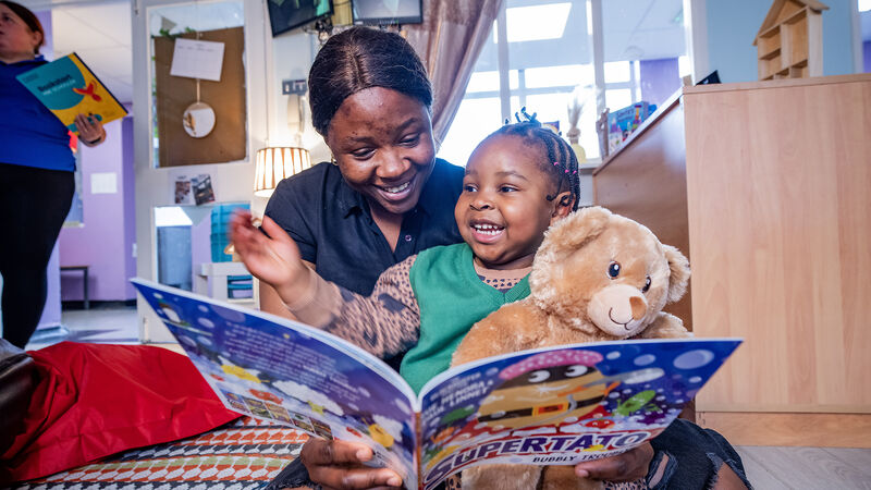 A woman and child holiday a teddy bear, laughing while sharing a Supertato book from a Bookstart pack in South Belfast
