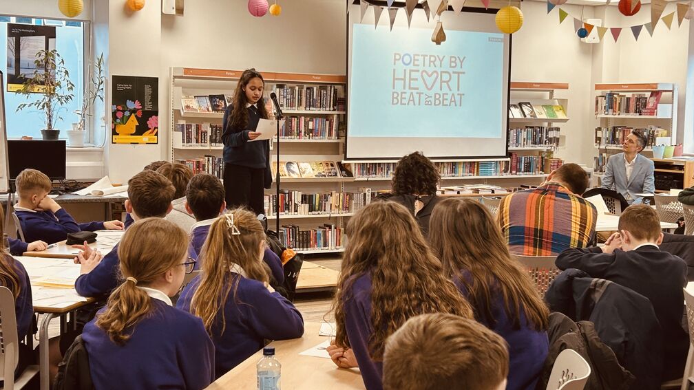 A classroom of children with a Poetry by Heart logo at the front as a child reads aloud