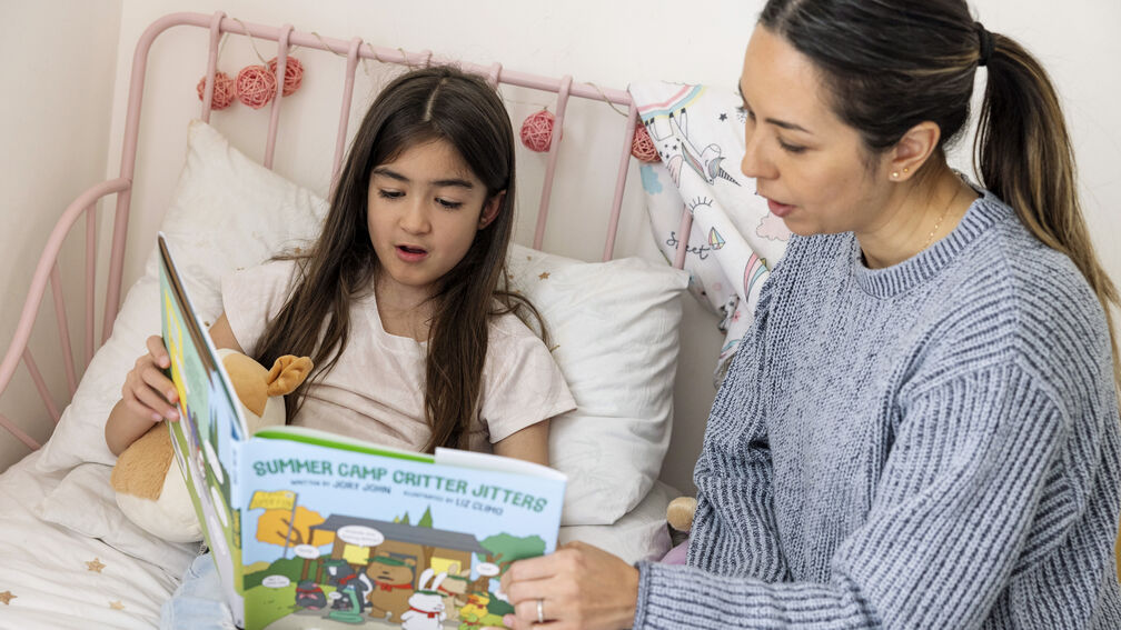 Young girl sitting on a bed next to carer looking down at a book and reading together