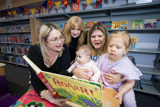 Librarian, mothers, and young children all reading a book together in the library