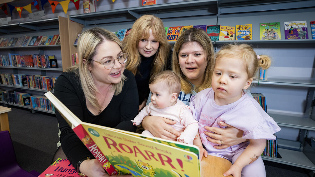 Librarian, mothers, and young children all reading a book together in the library