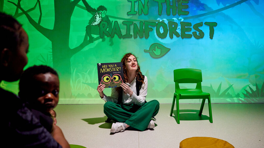 A woman sitting on a floor holding up the book Are You A Monster? during a story time session - behind her is a rainforest scene on the wall
