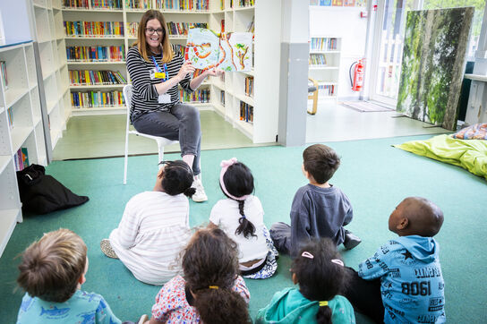 A femaie teacher is reading a picture book to a group of small children sitting on the floor in front of her