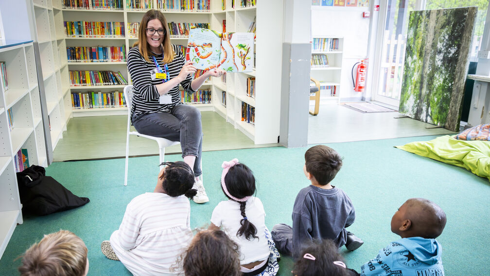 A femaie teacher is reading a picture book to a group of small children sitting on the floor in front of her