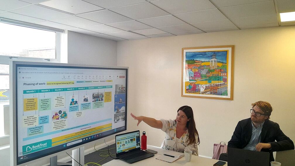 two people sitting down at a table, looking up at a screen. Person on the left is pointing their finger towards the screen.