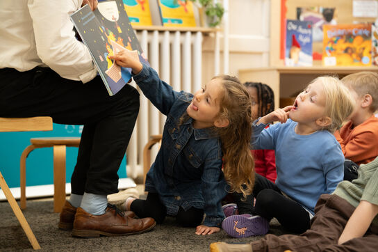 children sitting on the floor, the girl sitting at the front points towards the picture book being held up by an adult