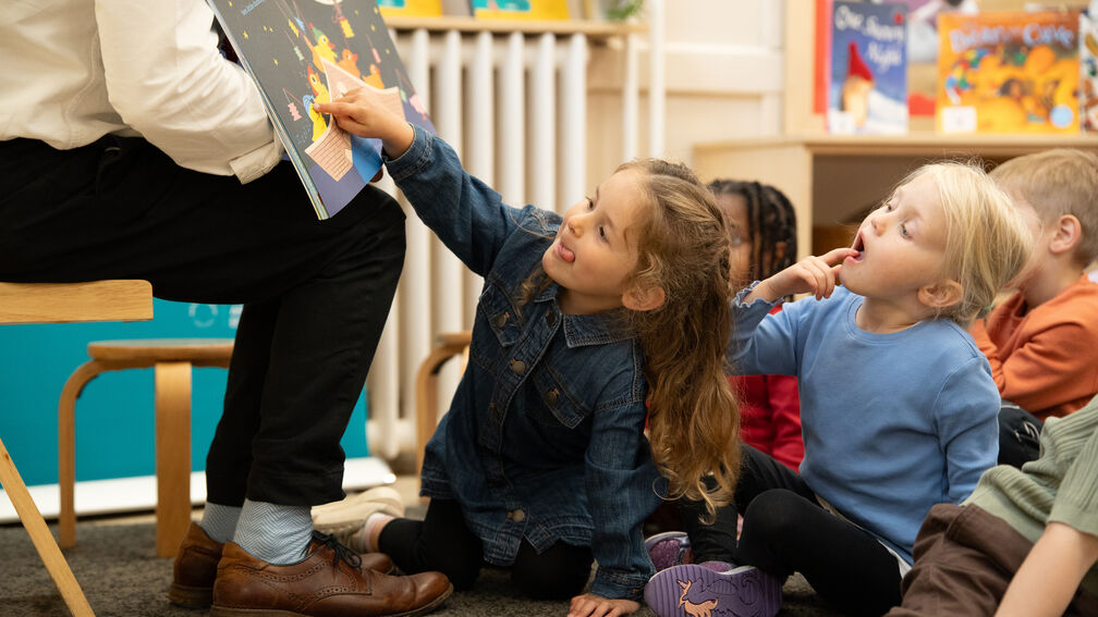 children sitting on the floor, the girl sitting at the front points towards the picture book being held up by an adult