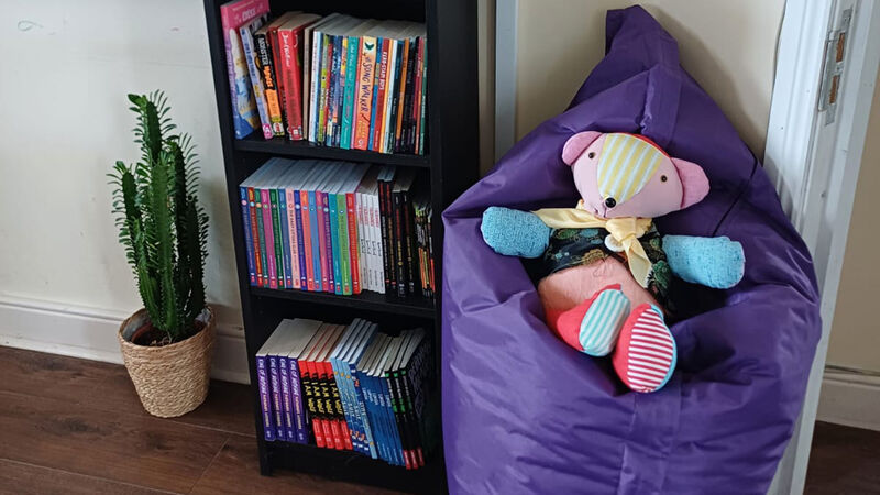 A reading area including a plant, a bookcase full of books, and a beanbag with a teddy on it