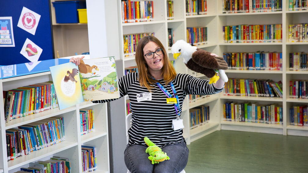 Person (Lucy Rowland) sitting on a chair face on holding up a picture book and an eagle puppet