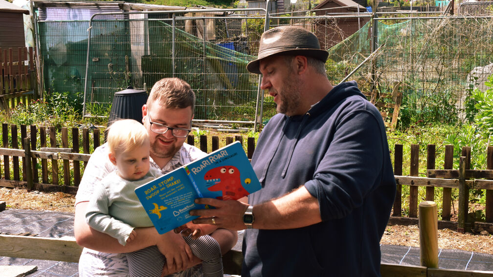 person standing and reading a book to a parent and child sitting down