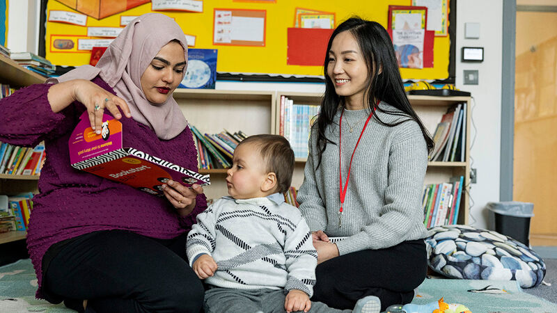A woman opening a Bookstart Baby pack, watched by a baby and a smiling woman wearing a lanyard