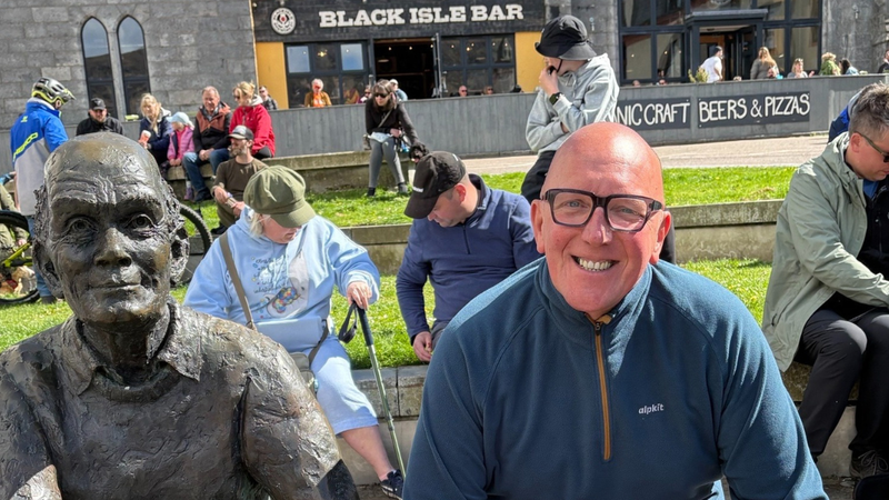 man smiling and sitting on bench outside next to a statue of a person