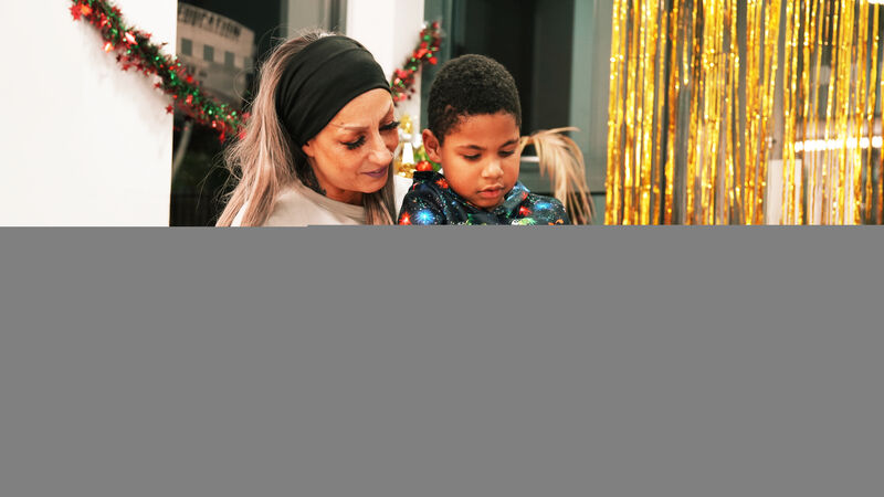 A mother with long blonde hair sat with her son on her lap, reading a book together. The room is decorated for Christmas.