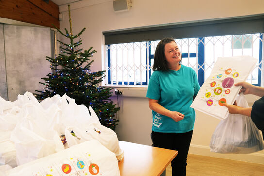 A woman in front of a table piled with food parcels, handing a BookTrust Christmas bookpack to someone not pictured