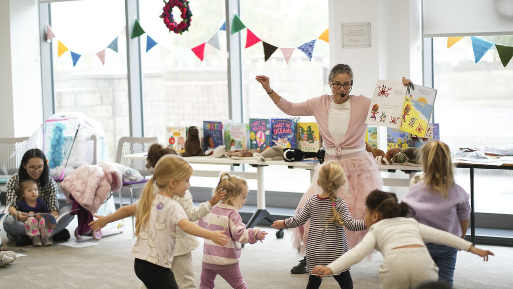 A woman in a pink outfit and a microphone headset is dancing with some young children in a library setting while holding up a picture book