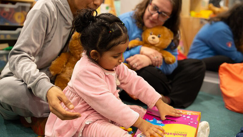 A child opening a Bookstart pack as a nursery worker watches on cuddling a reading bear