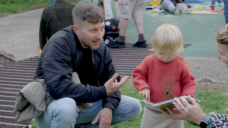 Dad sitting down with toddler son who is reading a book