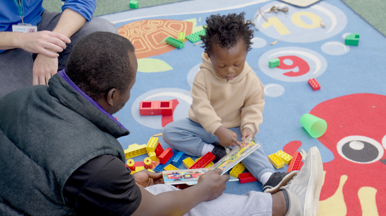 dad and son sitting on a colourful play-mat with lego reading a book