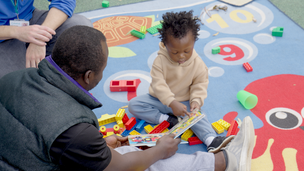 dad and son sitting on a colourful play-mat with lego reading a book