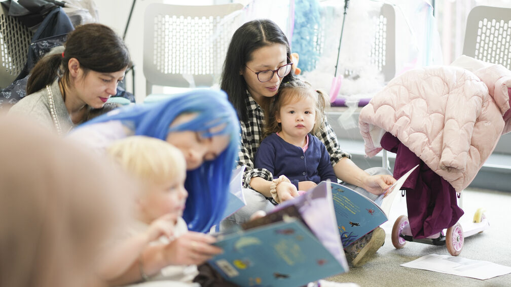 A group of mums is sitting on the floor of a library with their children in their laps, all reading a picturebook together.