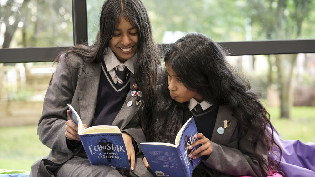 Two secondary school aged girls are sitting together in a school library setting and reading a book