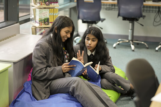 Two girls in school uniforms are sitting on beanbags in a school library, both reading the same book and having a fun discussion about it.