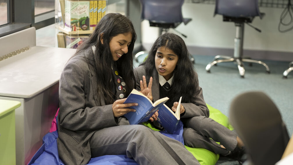 Two girls in school uniforms are sitting on beanbags in a school library, both reading the same book and having a fun discussion about it.