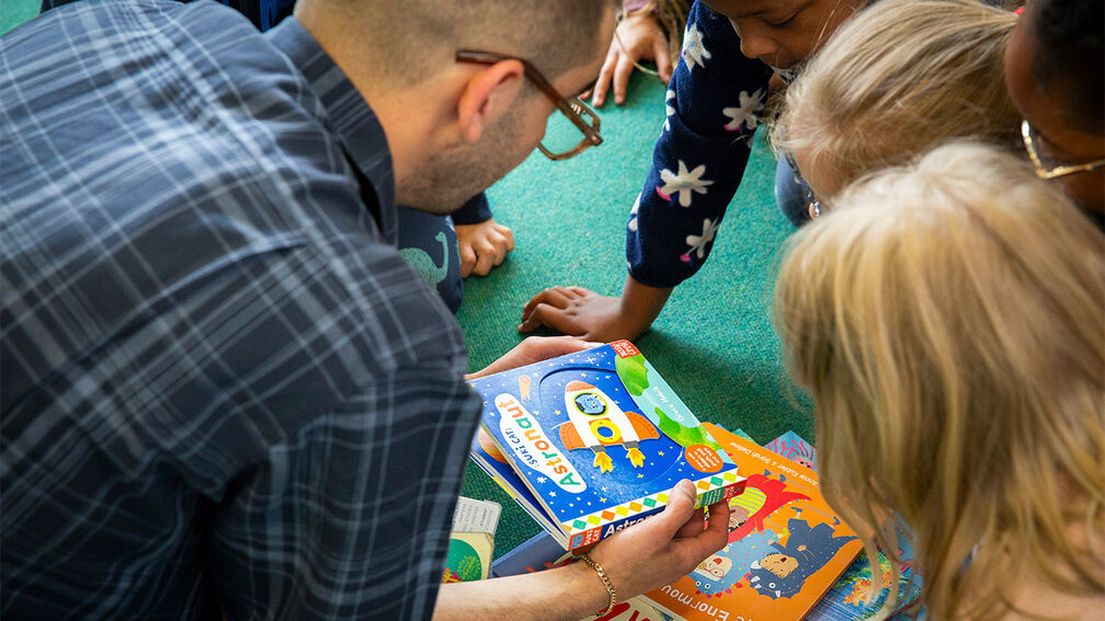 A teacher and children on the floor together - the teacher is showing the children a book with more books in a pile below
