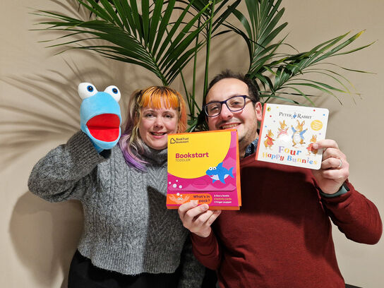 two people facing the camera and posing at the camera holding up a puppet and books
