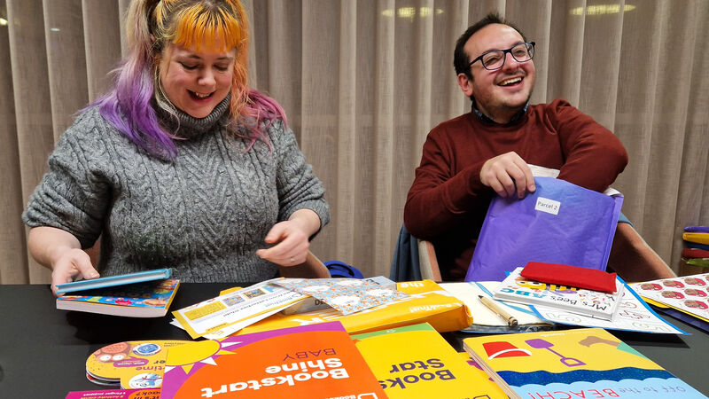 two people sitting at a table smiling with colourful books on the table