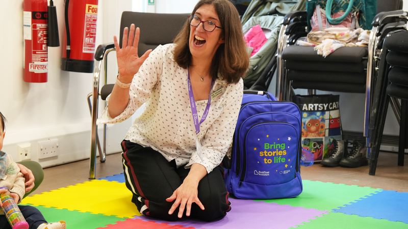 A white woman with brown hair and glasses is smiling and waving while sat on the floor in a library setting, next to her is a purple Bookstart Storyteller backpack, which has "Bring stories to life" embroidered on it.
