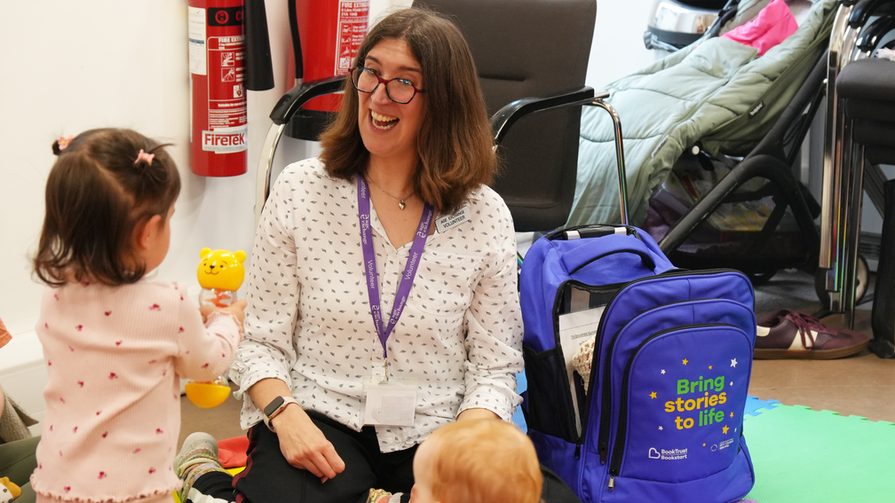 A white woman with brown shoulder length hair is smiling and interacting with a toddler in a library setting.