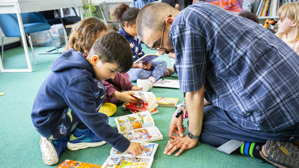 A young primary school aged boy is sat on the floor with his teacher, they are both looking at a book together