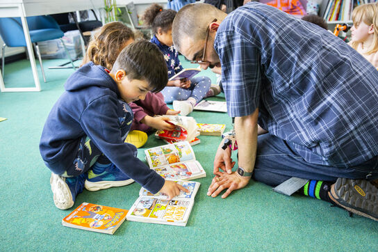 A young primary school aged boy is sat on the floor with his teacher, they are both looking at a book together