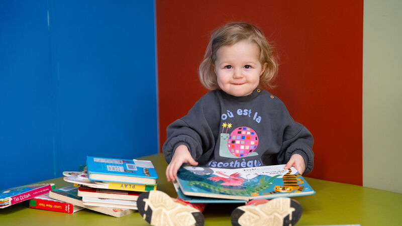 A toddler is sat with a big pile of books, reading and smiling at the camera
