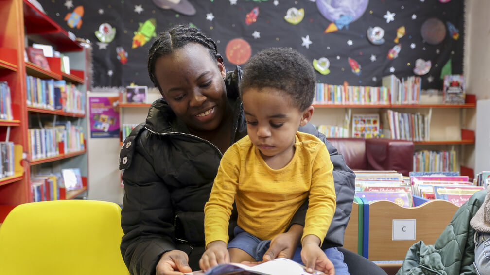 A boy is sat on the lap of his mother, they are both looking at the same picture book, in a library setting.
