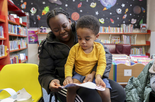 A boy is sat on the lap of his mother, they are both looking at the same picture book, in a library setting.
