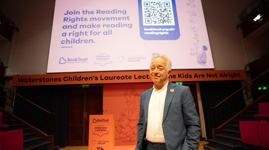 Frank Cottrell-Boyce standing on stage with large screen behind him with text saying 'Join the Reading Rights movement and make reading a right for all children.