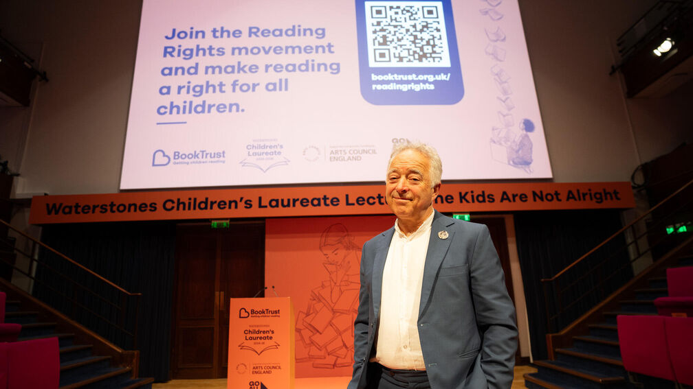Frank Cottrell-Boyce standing on stage with large screen behind him with text saying 'Join the Reading Rights movement and make reading a right for all children.